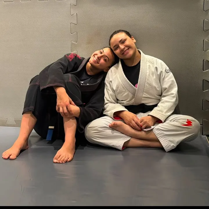Monique Fernanda, de kimono branco, sentada no tatame ao lado de uma aluna